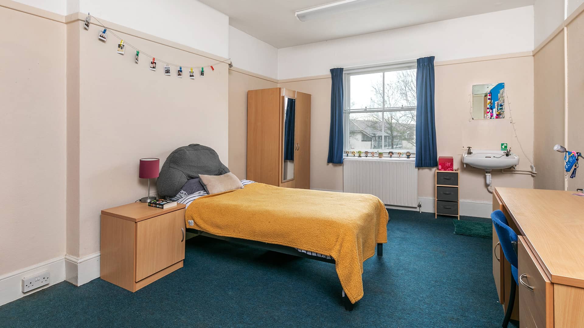 A dorm room with a single bed, a wooden desk and chair, a wooden wardrobe, and a sink with a mirror above it