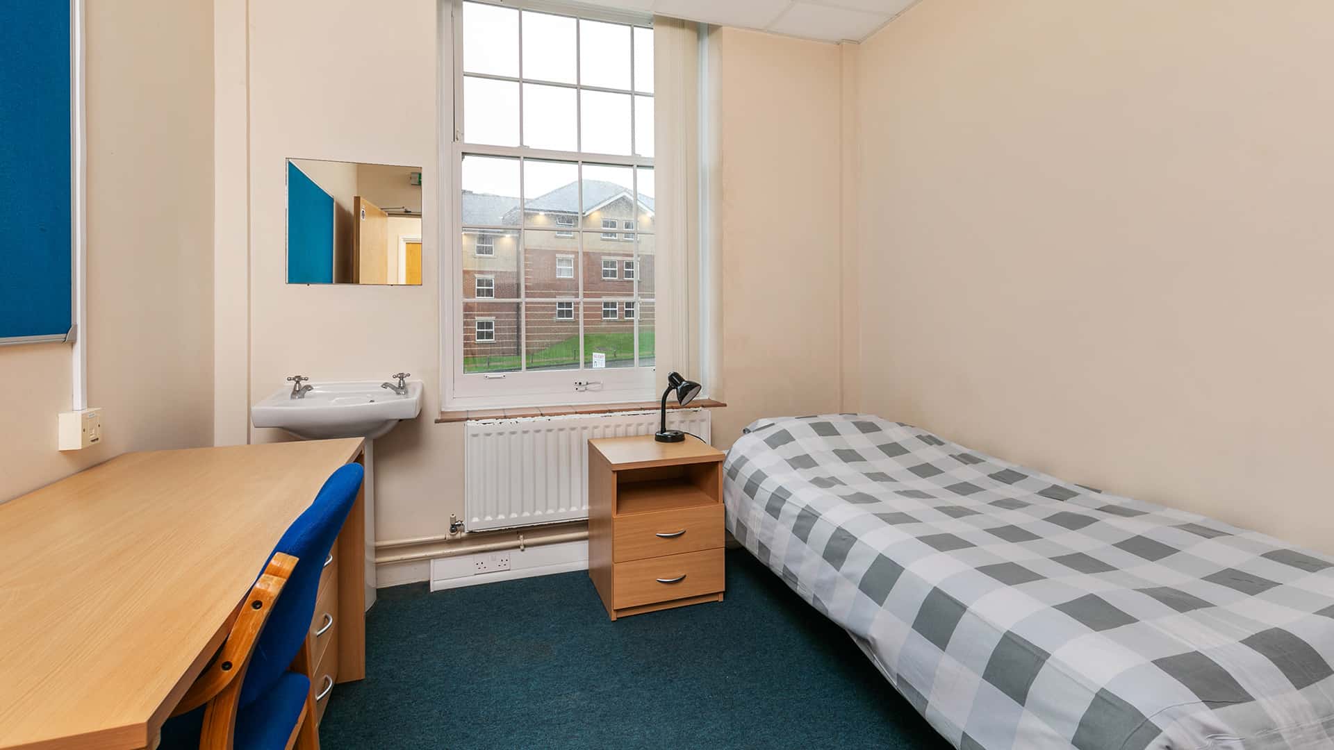 A dorm room with a single bed, a wooden desk with a chair, a small bedside table with a lamp, and a window with a view of a brick building