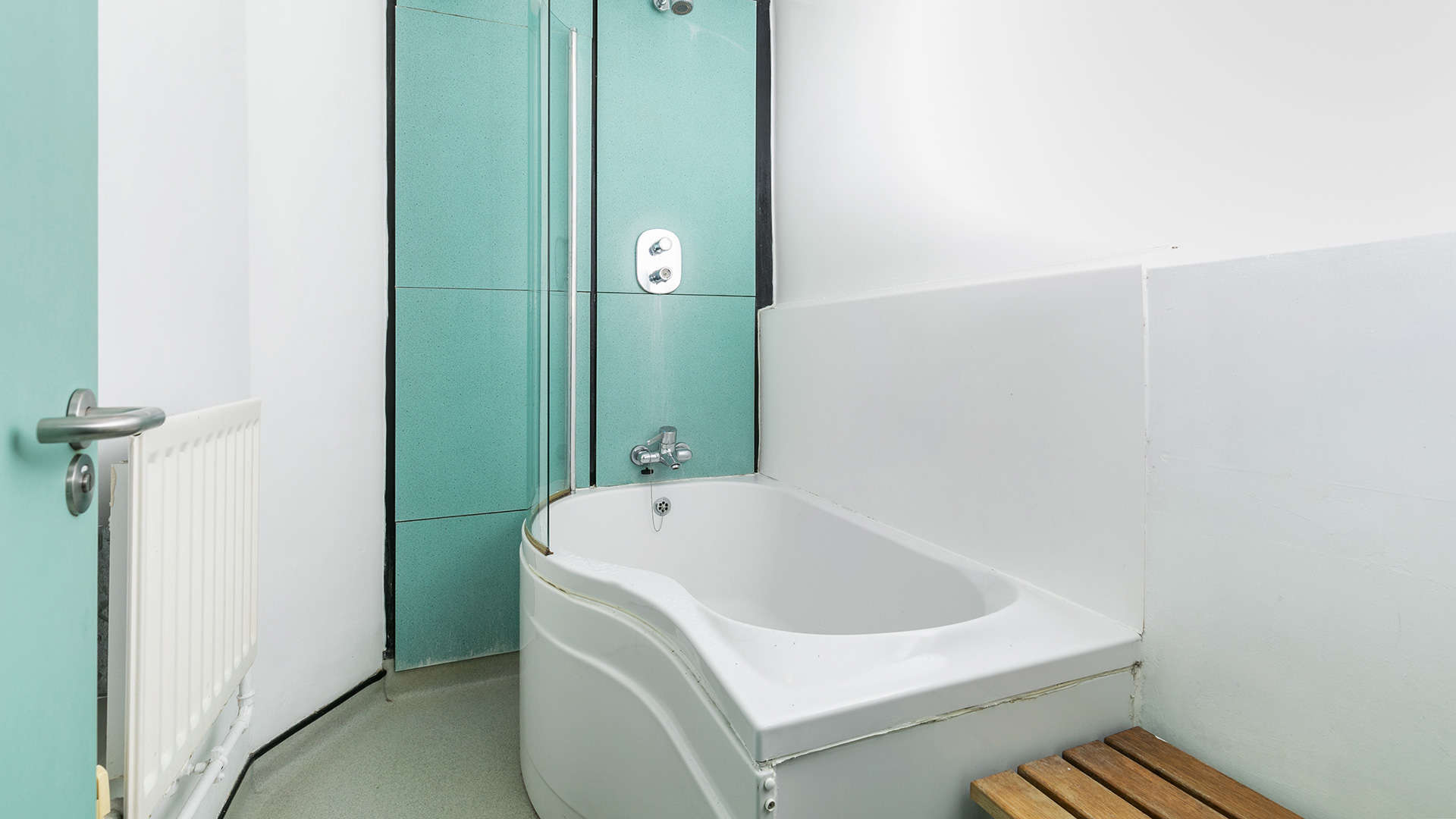 A bathroom with a corner bathtub and shower combo with a glass partition, a wooden bath mat, and a radiator.