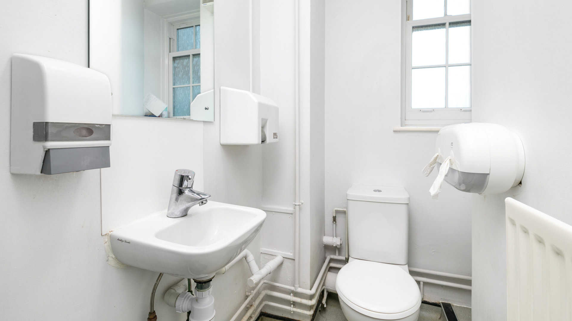 A bathroom with a toilet, sink, two paper towel dispensers, and a small window with frosted glass.