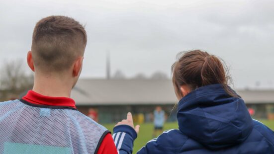 Student coaching a school pupil on the touchline of a football pitch