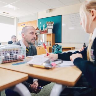 A teaching student crouched over a desk to talk to a pupil