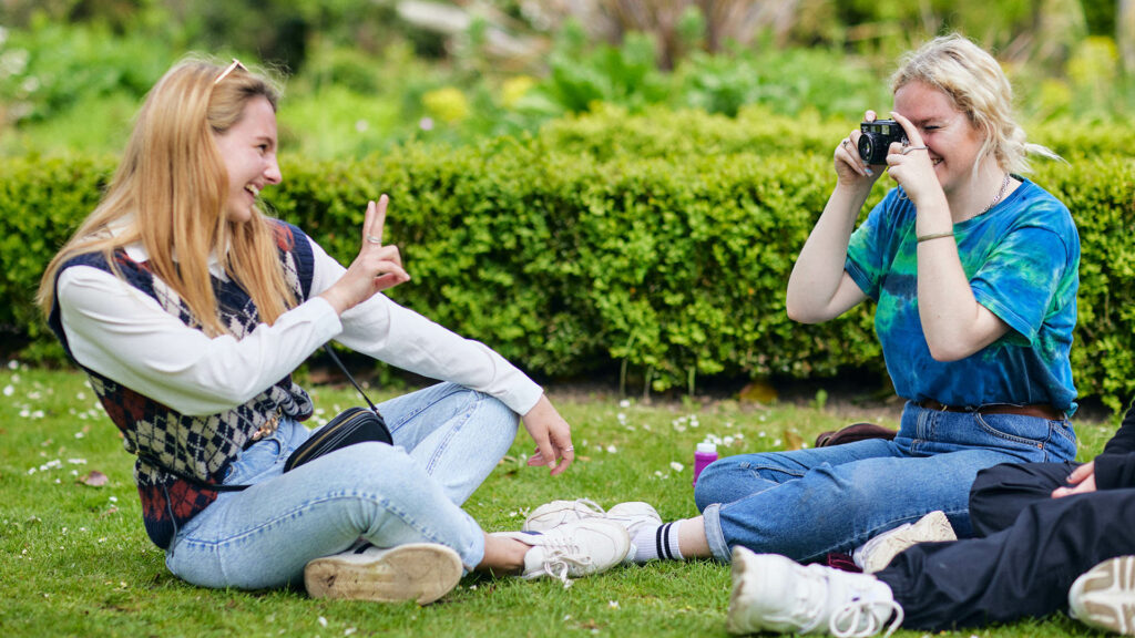 21 Students Chichester Park 1920 X 1080 5 students sitting on grass laughing and taking pictures of eachother
