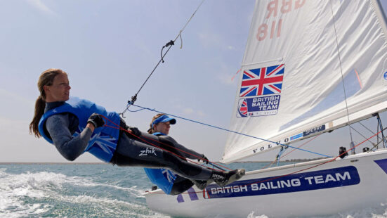 A female sailor hanging off the edge of a professional sailing boat during a competition.