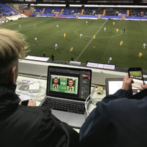 Sports Media students working on the social media for a football match as the game continues below them on the pitch.