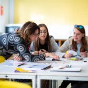 Teaching tutor leaning across a desk to help two students with their work