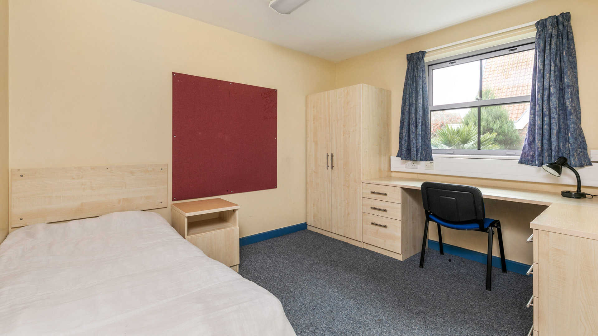 A simple bedroom with a single bed, wooden furniture including a wardrobe, desk, and bedside table