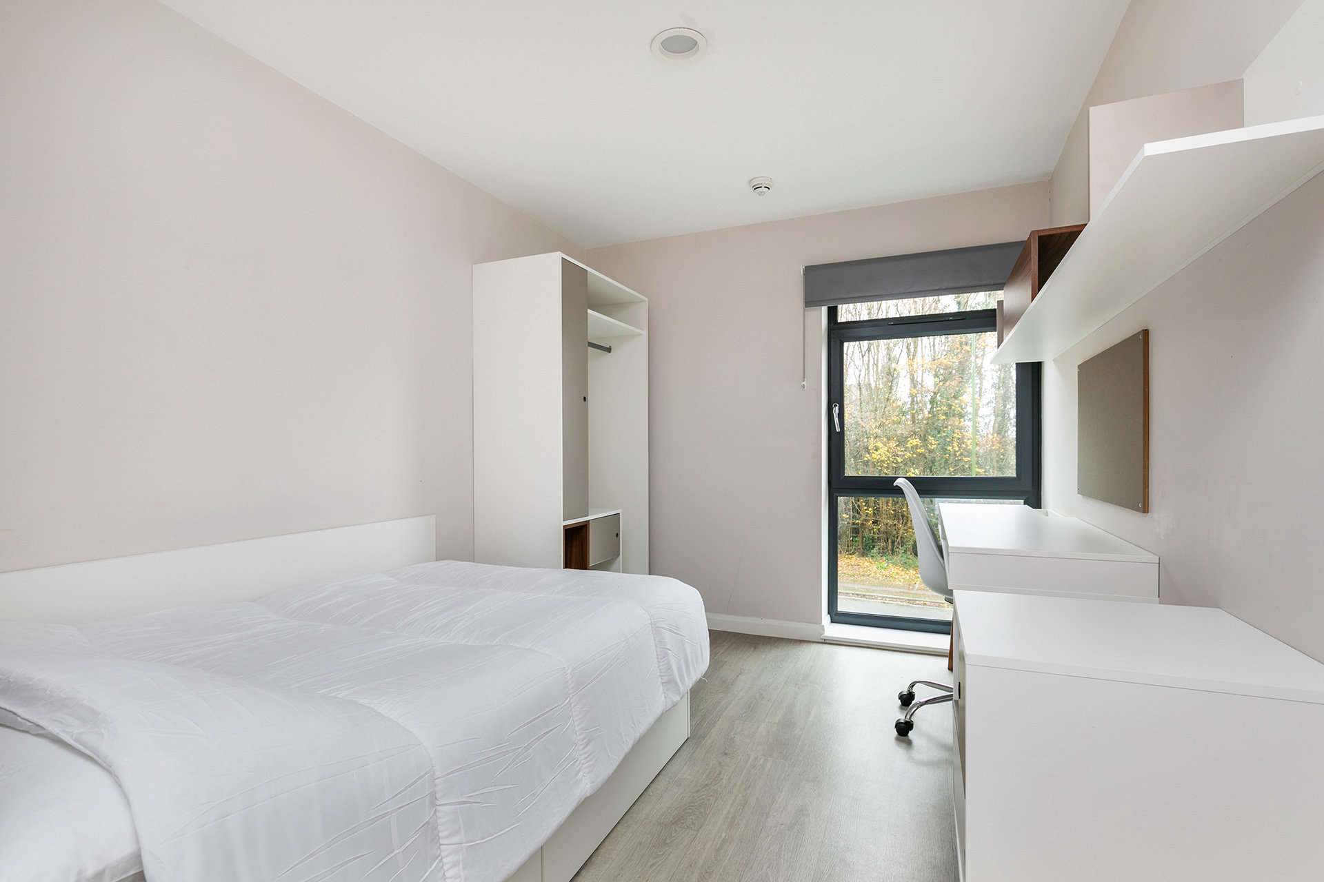 A modern bedroom with a single bed, a built-in closet, and a desk with a chair by a large window overlooking a wooded area.