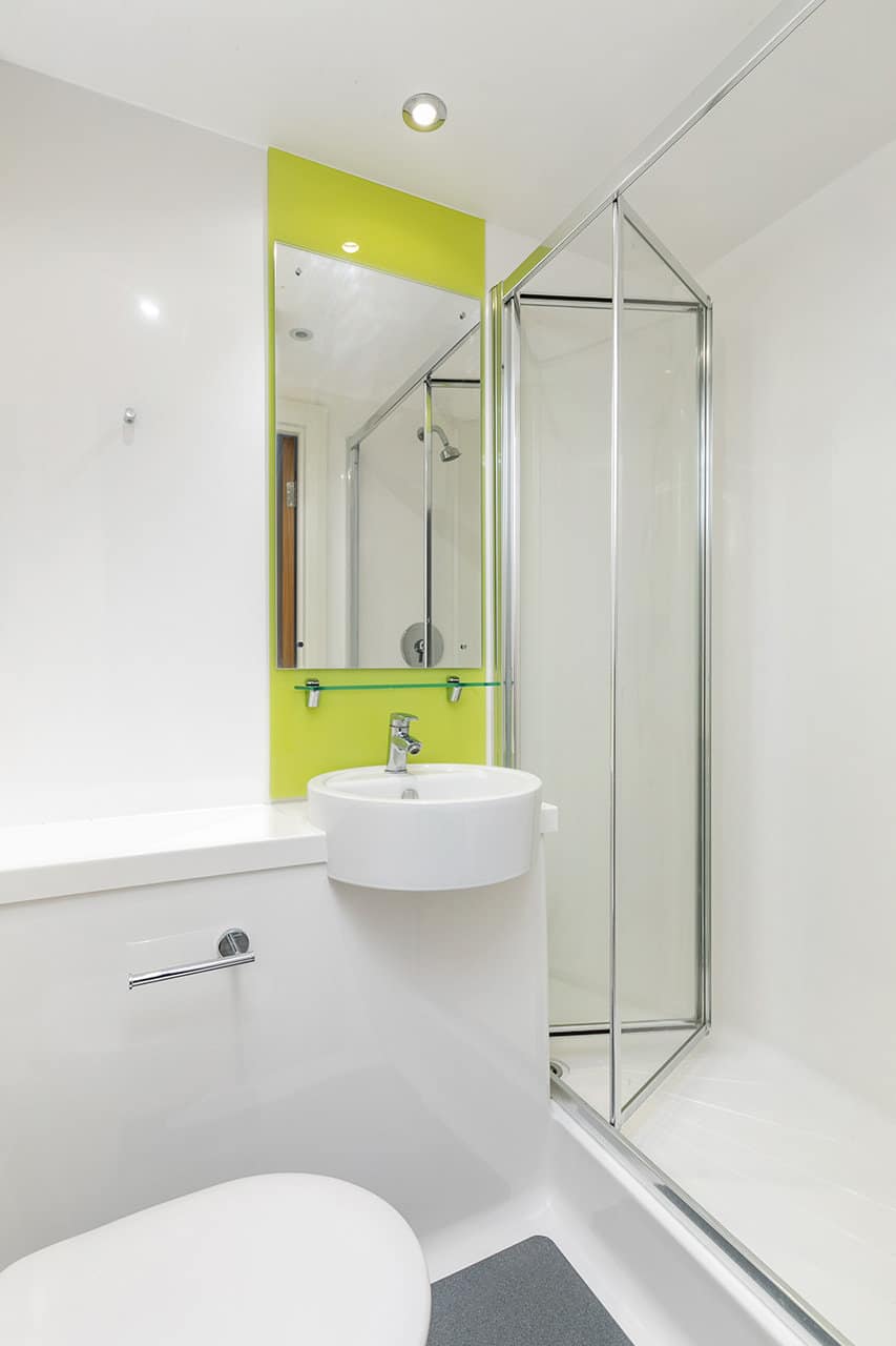 A modern bathroom with a white bathtub, a round white basin, and a glass shower door with a lime green accent wall and mirror above the sink.