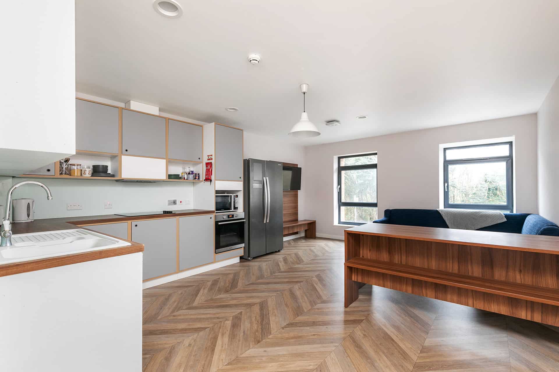 Modern kitchen with light gray cabinets, wooden countertop, stainless steel appliances and an attached dining area with a long wooden bench and dark blue cushions