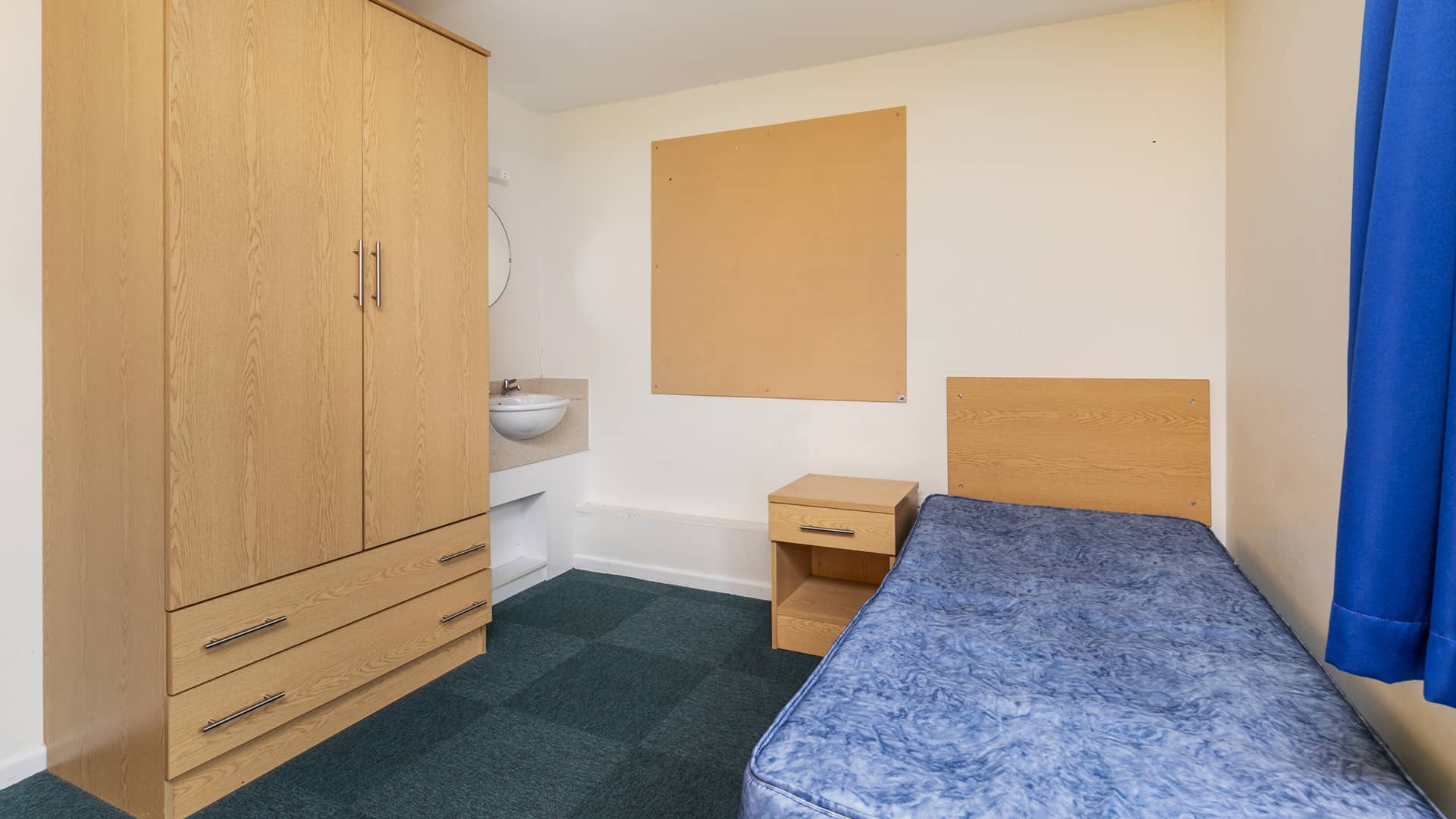 A dormitory room with a wardrobe, single bed, a sink, and a cork board on the wall.