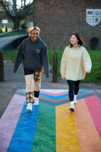 Two young students are walking across a Pride flag painted on the ground on campus